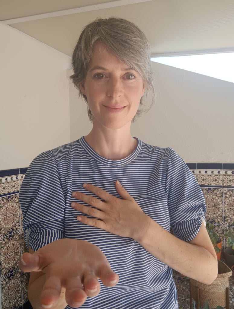 Catelijne Coopmans looking at the camera, one hand on her heart and the other with palm upwards reaching forward. A gesture of connection inwards and outwards. 